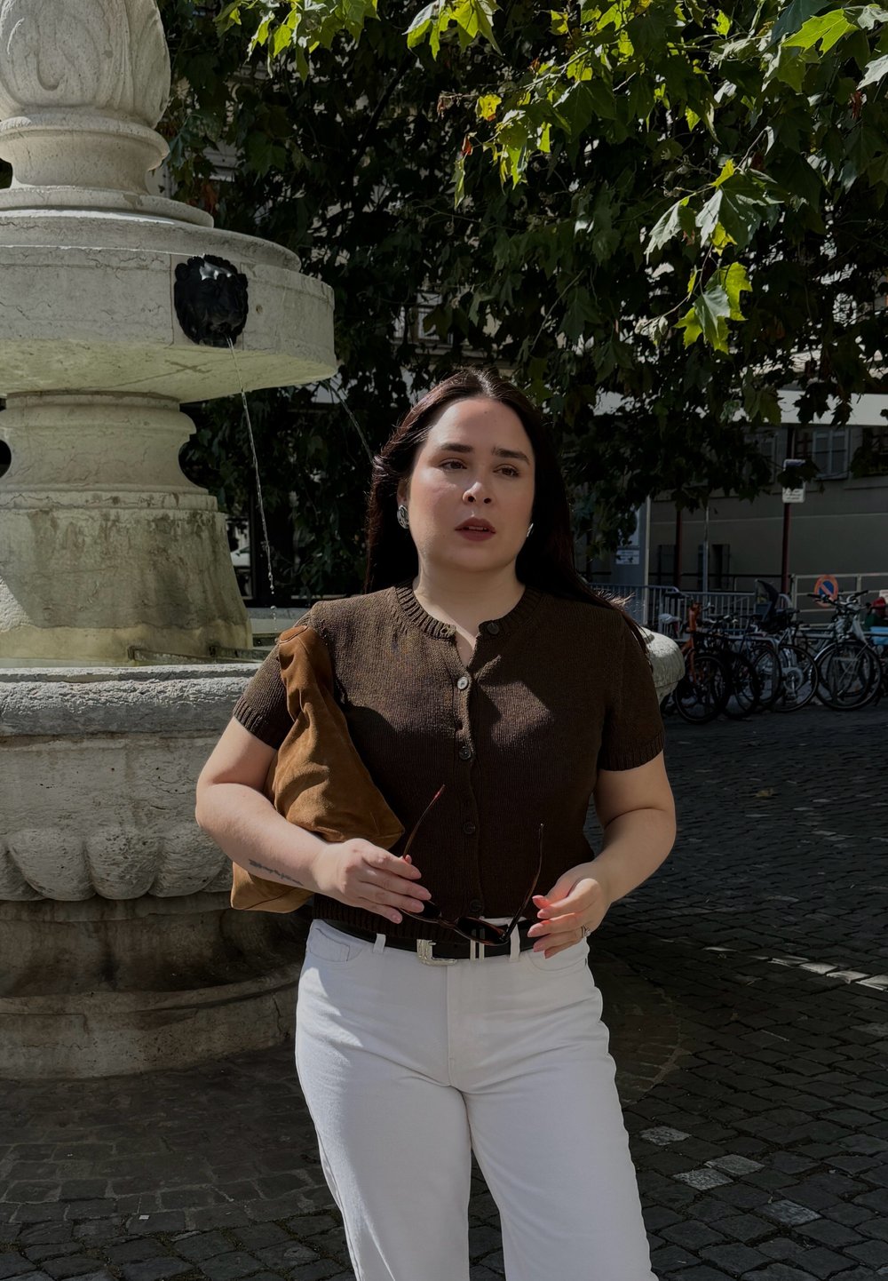 Bruine gebreide cardigan met korte mouwen, witte hoge taillebroek, houdt een zonnebril vast, met een bruine suède schoudertas, staat naast een stenen fontein.