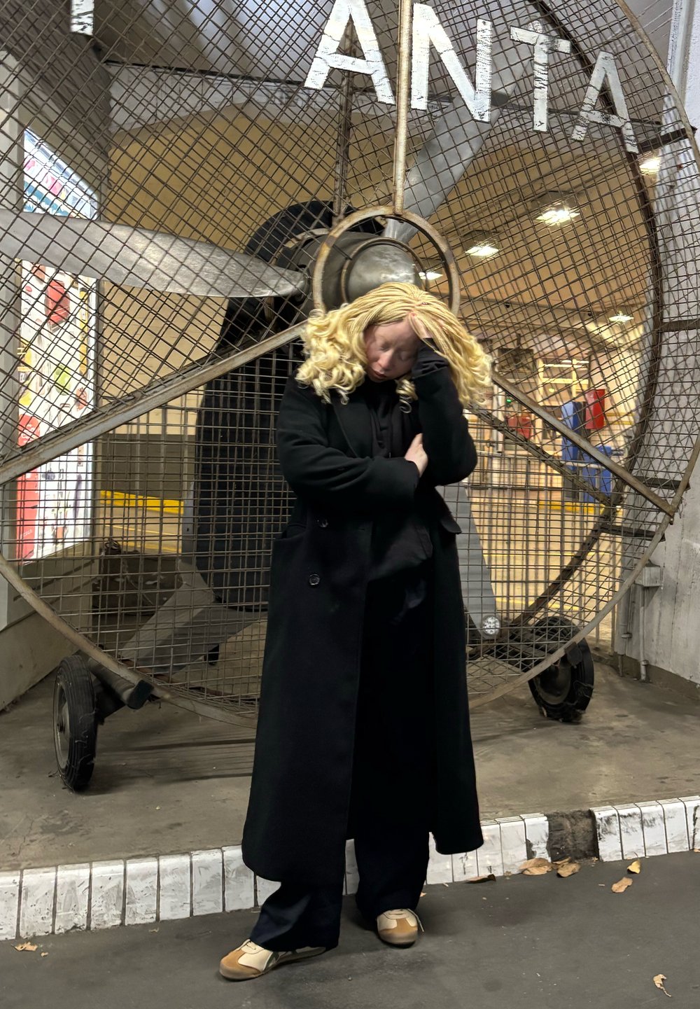 Un manteau noir et un pantalon foncé sont portés avec des chaussures de couleur claire. Un grand ventilateur en métal sert de fond, présentant un motif en grille et de la rouille.