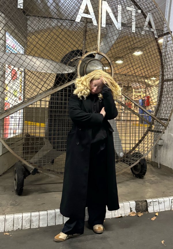 Crni kaput i tamne hlače nose se s svijetlim cipelama. Veliki metalni ventilator služi kao pozadina, s uzorkom rešetke i hrđom.