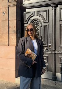 Szary, oversize blezer, biały top, jasnoszare spodnie, trzymający magazyn "Vogue". Czarne okulary przeciwsłoneczne. Teksturowane czarne drewniane drzwi w tle.