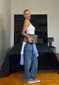 Femme en haut blanc sans manches et jean bleu ample tenant une pochette imprimée léopard, debout à côté d'un lit noir avec une lampe sur un sol en bois.