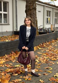 Modella che indossa un blazer a righe blu navy, una t-shirt grafica, pantaloni corti neri, collant trasparenti e mocassini scuri, mentre tiene in mano una borsa intrecciata color bordeaux.