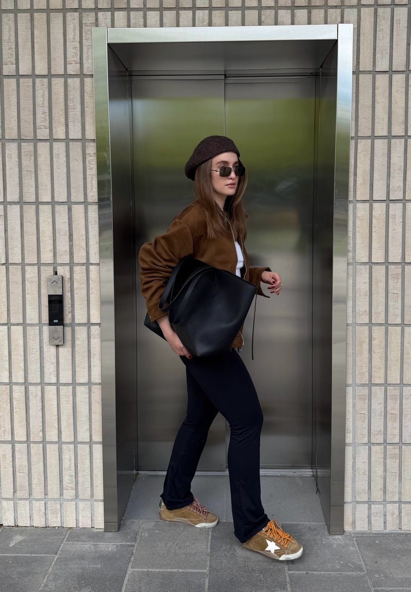 Femme portant des lunettes de soleil et une veste marron, portant un grand sac noir, entrant dans un ascenseur en acier inoxydable dans un bâtiment carrelé.