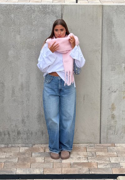 Bufanda rosa clara, camisa blanca oversized, jeans azules holgados y calzado marrón. De pie contra un fondo de concreto gris.