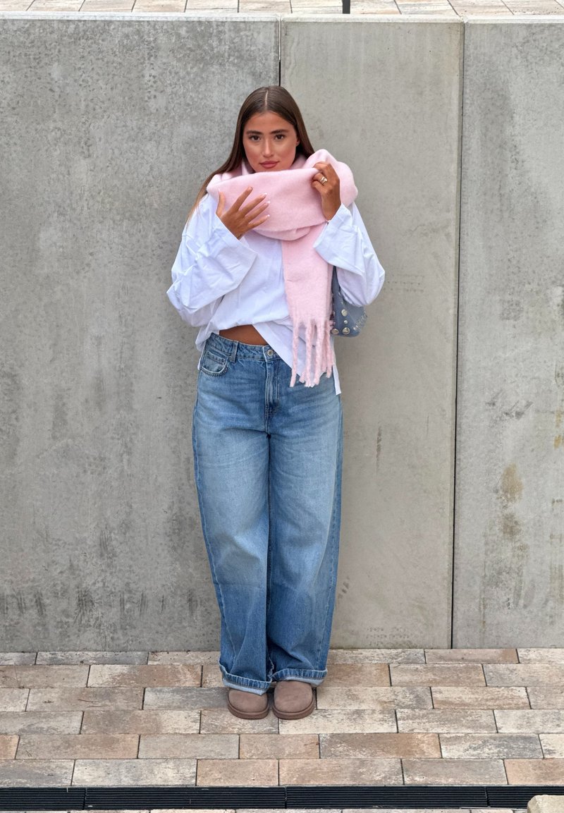 Cachecol rosa claro, camisa branca oversized, jeans azul folgados e calçado castanho. De pé contra um fundo de betão cinza.