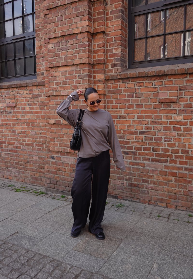 Sieviete ar saulesbrillēm, pelēkā garroku krekla un melnām biksēm stāv un smaida pie sarkanas ķieģeļu sienas ar lieliem logiem.