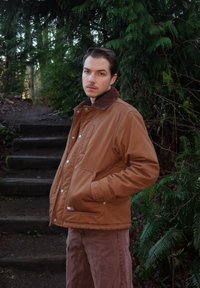 Veste isolante marron avec col en velours côtelé, boutons-pression et poches latérales ; portée avec un pantalon marron clair ; entourée d'un feuillage vert luxuriant.