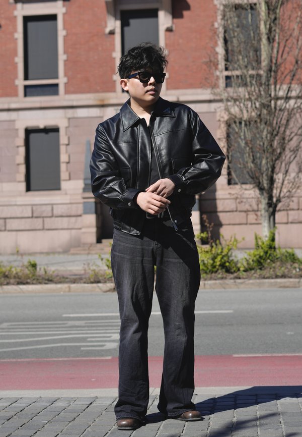 Osoba ubrana w czarną skórzaną kurtkę, ciemne dżinsy, okulary przeciwsłoneczne, stoi na chodniku przed ceglaną budynkiem z oknami i drzewami bez liści.