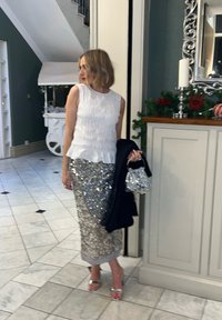Haut blanc sans manches avec un ourlet peplum, associé à une jupe crayon argentée à sequins. Tenant un petit sac à main argenté ; portant des talons argentés.