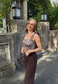 Leopardenmuster-Top aus glattem Stoff, kombiniert mit hoch geschnittenen, braunen, strukturierten Hosen; steht vor einer Steinwand.