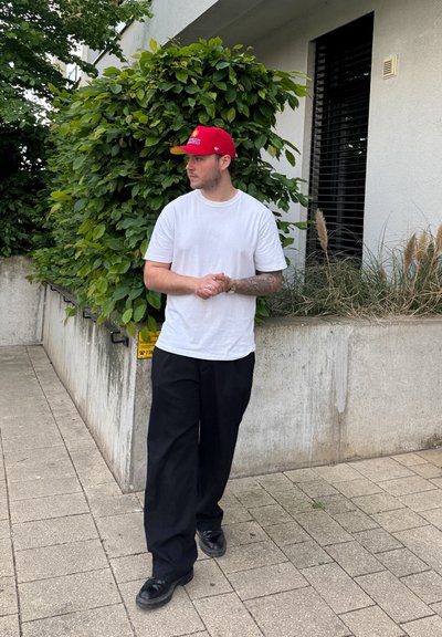 Camiseta blanca, pantalones de pierna ancha negros y zapatos negros. Gorra de béisbol roja con acentos amarillos, de pie junto a un arbusto verde y una pared de concreto.