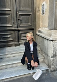 Blazer oversize nero, camicia bianca e mocassini neri chunky indossati con una gonna corta. Tiene una piccola borsa bianca sulle scale in pietra accanto a una porta scura.