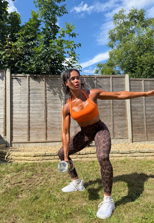 Mujer con sujetador deportivo naranja y leggings con estampado de leopardo hace ejercicio al aire libre con una mancuerna, estirando el brazo hacia un lado en un jardín bajo el cielo azul.