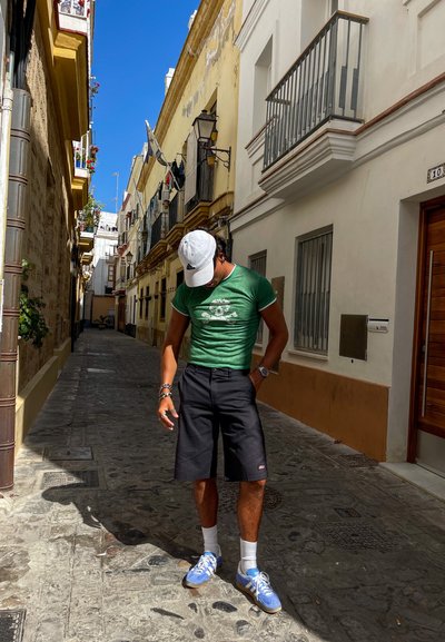 Camiseta verde ajustada con un diseño gráfico, pantalones cortos negros, calcetines blancos y zapatillas azules. Entorno urbano informal con pavimento de adoquines.