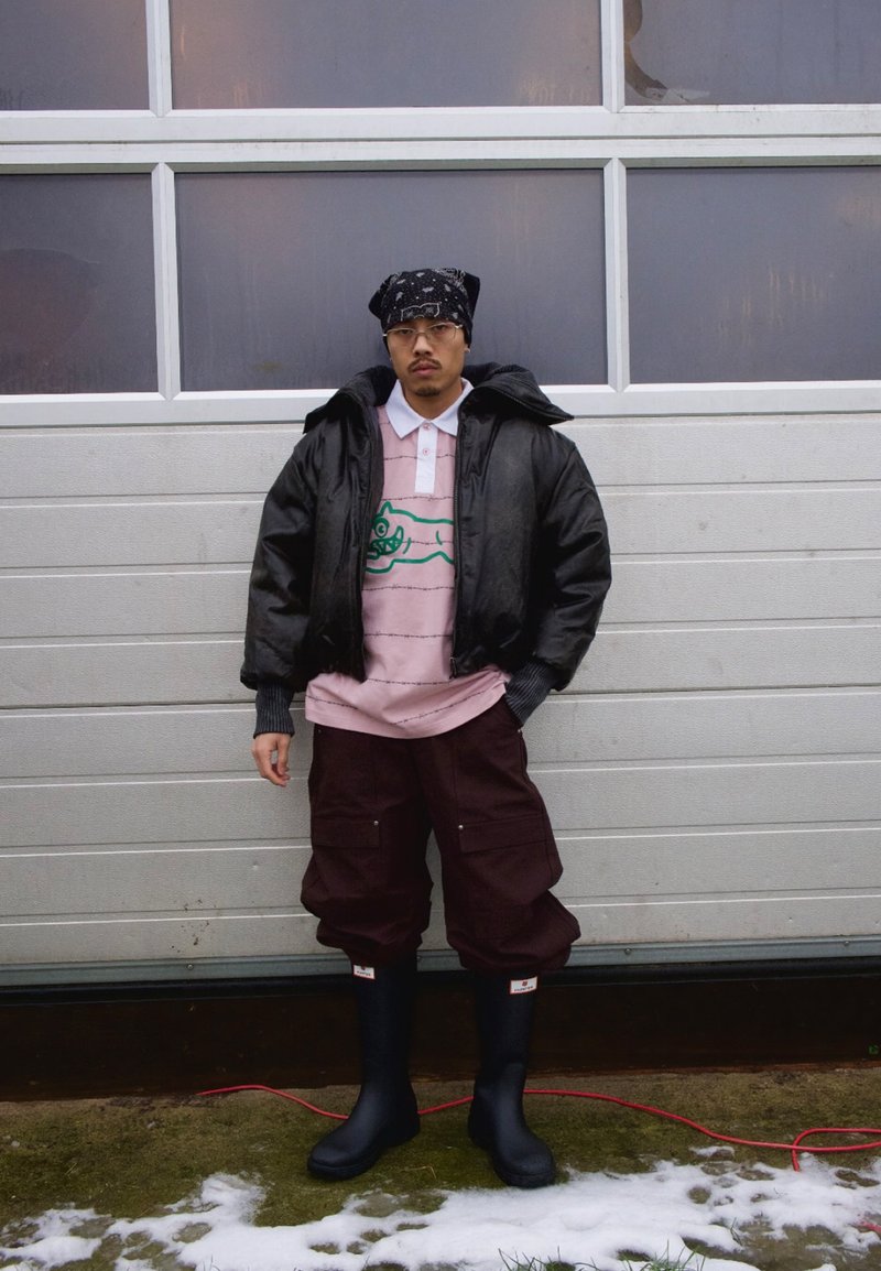 Man som bär svart vadderad jacka, rosa pikétröja med tryck, mörka cargobyxor, svarta gummistövlar och svart bandana, stående på snötäckt gräs.