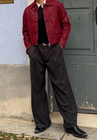 Άτομο που φοράει κόκκινο δερμάτινο μπουφάν, μαύρο παντελόνι με φαρδιά μπατζάκια, μαύρα παπούτσια, στέκεται με τα χέρια στις τσέπες μπροστά σε γκρι και μπλε τοίχους.