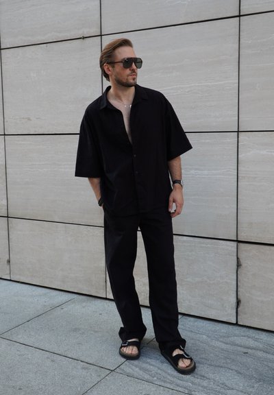 Camisa negra de manga corta con botones combinada con pantalones a juego. Lleva sandalias negras y gafas de sol oscuras, con un fondo de piedra clara.