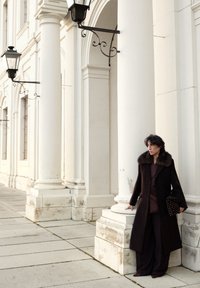 Personne en manteau foncé à col en fourrure tenant un sac à motifs, debout contre une colonne blanche d'un bâtiment classique avec des lanternes.