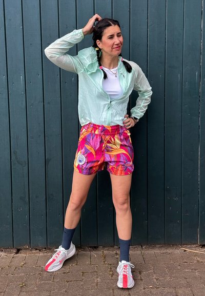 Chaqueta de manga larga, de color verde claro y con cremallera, sobre una camisa blanca; pantalones cortos rosa vibrante con un patrón de hojas; zapatillas blancas con rayas rojas.