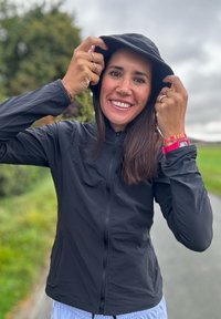 Black lightweight jacket featuring a high collar and zip fastening, paired with a matching black hat. It has long sleeves and a smooth texture, set in an outdoor environment.