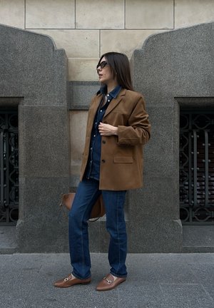 Blazer de ante marrón sobre una camisa de mezclilla, combinado con jeans azules y mocasines marrones. Los accesorios incluyen un bolso marrón y gafas de sol oscuras.