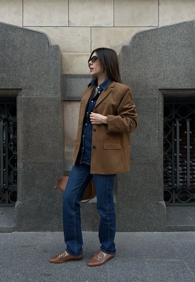 Blazer de ante marrón sobre una camisa de mezclilla, combinado con jeans azules y mocasines marrones. Los accesorios incluyen un bolso marrón y gafas de sol oscuras.