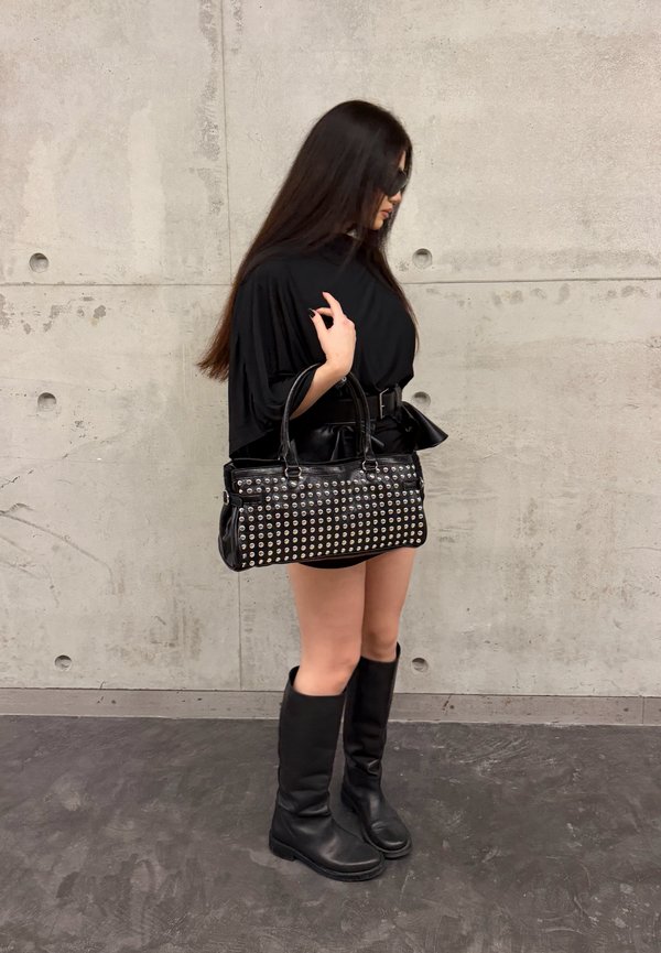 Une femme vêtue de noir et portant des bottes jusqu’aux genoux tient un sac à main noir clouté contre un mur en béton.