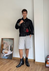 Czarna kurtka dżinsowa z dwiema kieszeniami na piersi, szare szorty sportowe, czarne skórzane botki za kostkę z dekoracyjnym przeszyciem oraz czerwona torba.