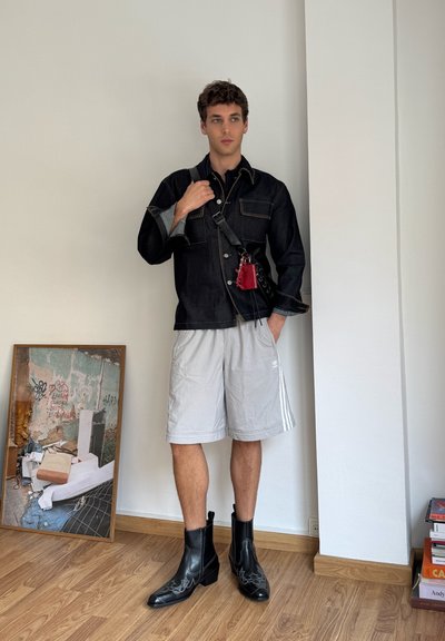 Chaqueta de mezclilla negra con dos bolsillos en el pecho, pantalones cortos deportivos grises, botas de cuero negras hasta el tobillo con costuras decorativas y una bolsa roja.