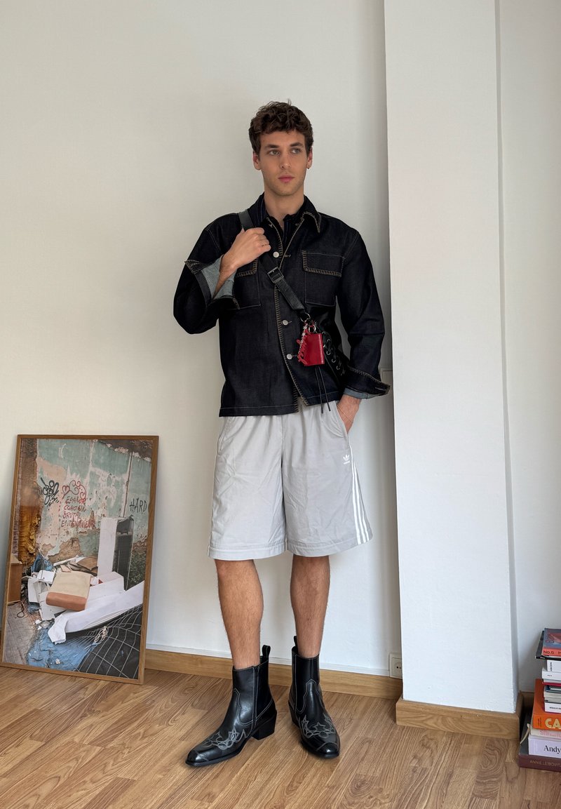 Giacca di jeans nera con due tasche sul petto, pantaloni corti sportivi grigi, stivali neri in pelle alla caviglia con cuciture decorative e una borsa rossa.
