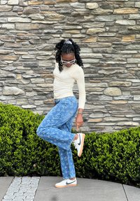 Uma blusa branca de mangas longas, jeans azuis com padrão floral e ténis Nike brancos com detalhes pretos. Fundo com uma sebe verde e uma parede de pedra.