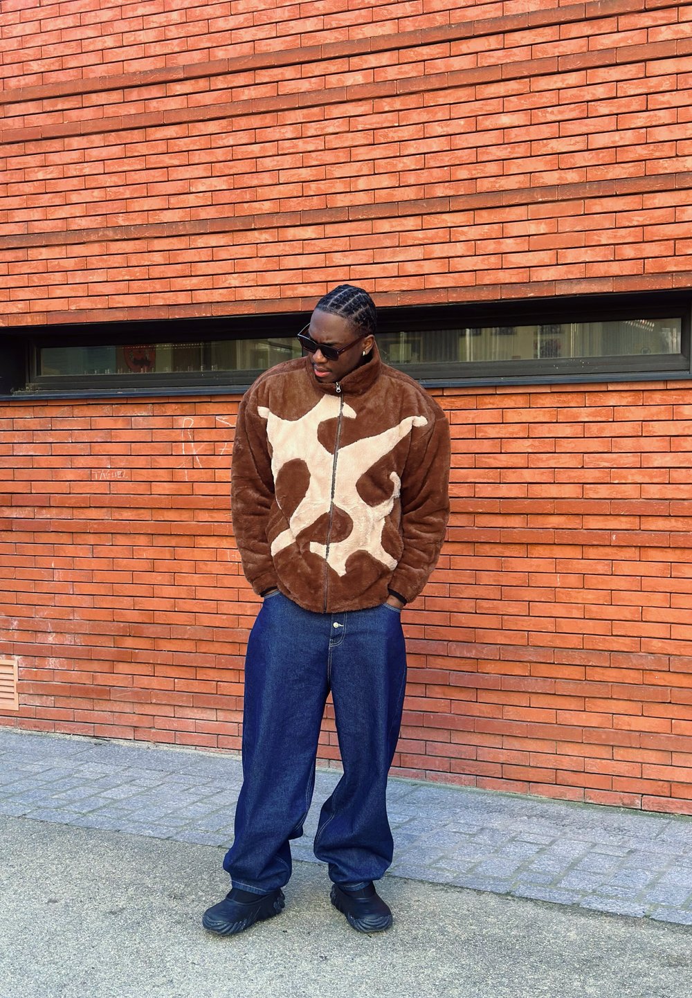 Veste en polaire marron avec un motif abstrait beige clair, fermeture éclair à l'avant, associée à un pantalon en denim bleu ample et des chaussures noires. Fond en brique.