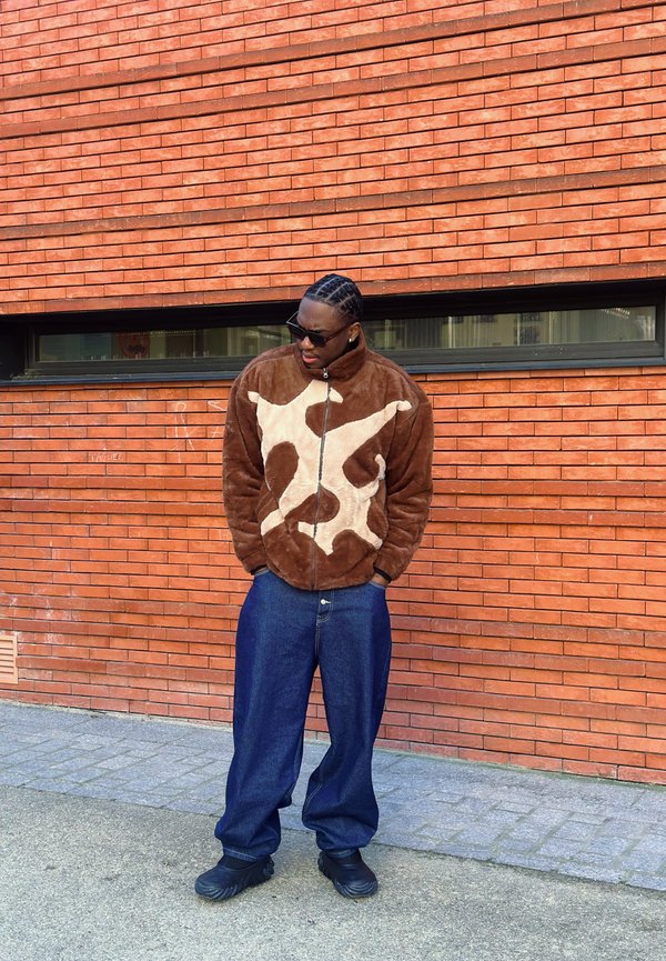Brown fleece jacket with a light beige abstract pattern, zip-up front, paired with loose blue denim trousers and black footwear. Brick wall background.