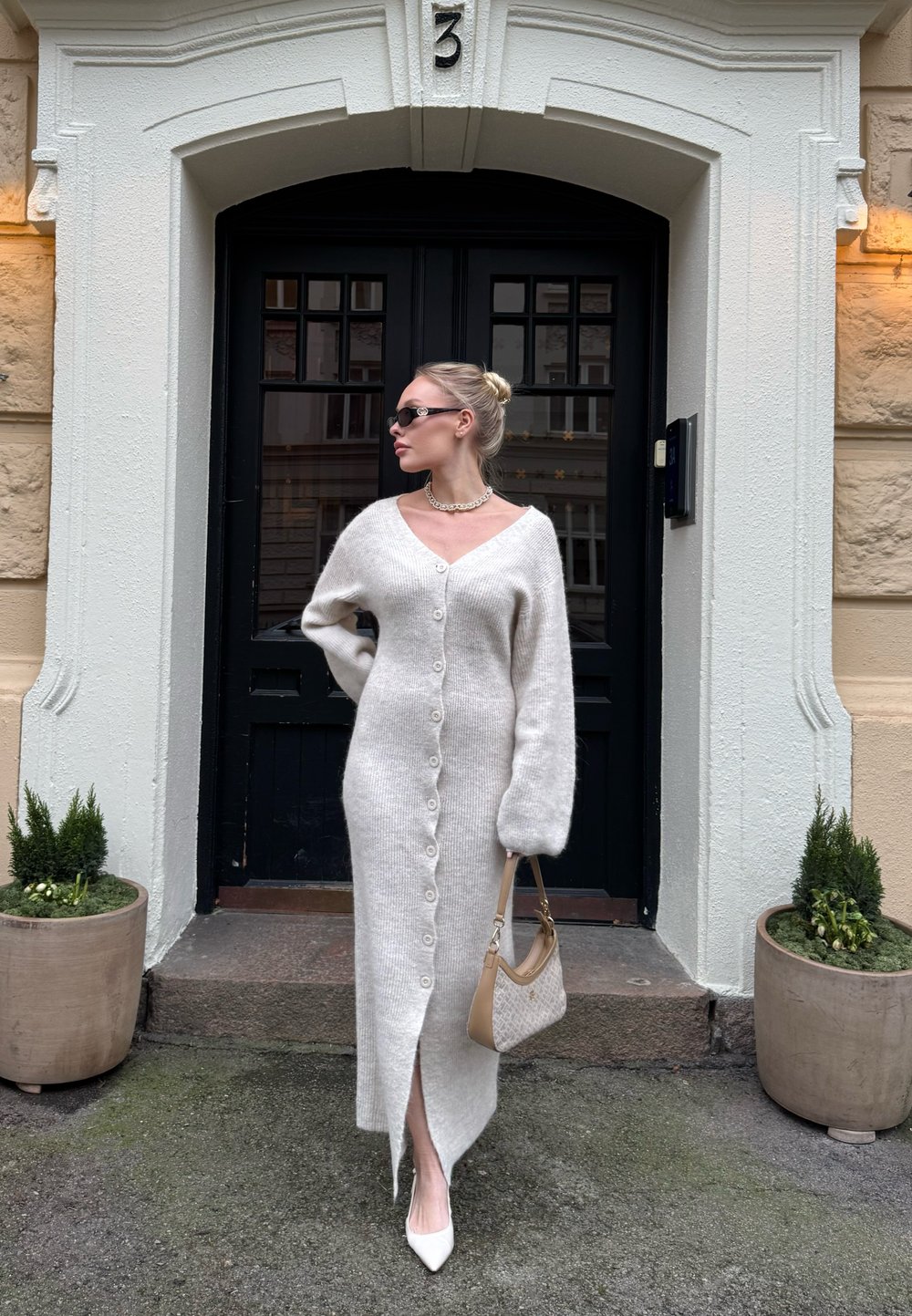 Γυναίκα με μακρύ λευκό πλεκτό φόρεμα με κουμπιά και λευκά τακούνια στέκεται με μια μπεζ τσάντα έξω από μια μαύρη πόρτα με τον αριθμό 3, πλαισιωμένη από γλάστρες με φυτά.