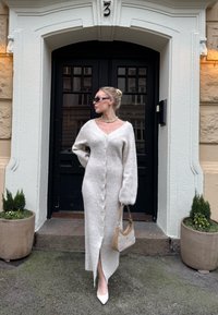 Γυναίκα με μακρύ λευκό πλεκτό φόρεμα με κουμπιά και λευκά τακούνια στέκεται με μια μπεζ τσάντα έξω από μια μαύρη πόρτα με τον αριθμό 3, πλαισιωμένη από γλάστρες με φυτά.