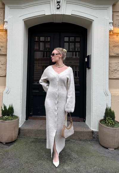 Mujer con un vestido largo de punto blanco abotonado y tacones blancos, de pie con un bolso beige frente a una puerta negra con el número 3, flanqueada por macetas.