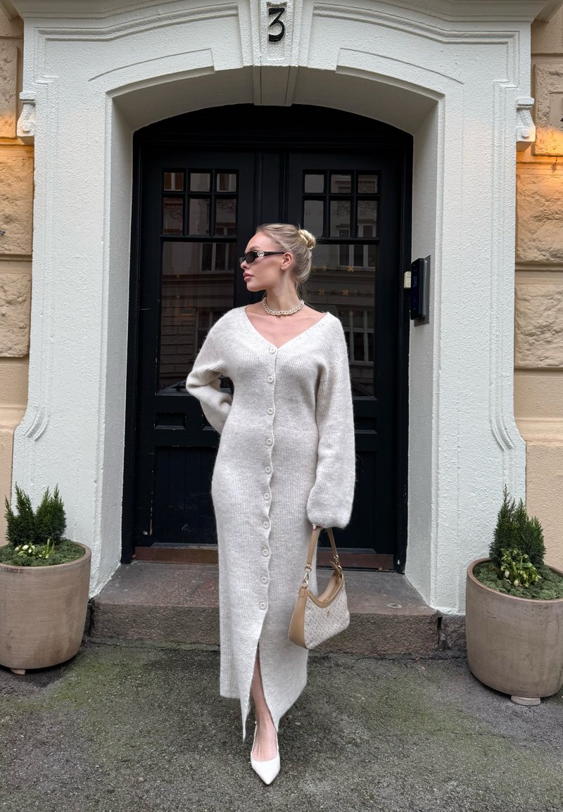 Kvinne i en lang hvit knappet strikkekjole og hvite hæler står med en beige håndveske utenfor en svart dør merket med nummer 3, flankert av plantepotter.