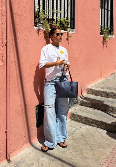 Mujer con gafas de sol, camiseta blanca y vaqueros anchos rasgados, de pie contra una pared rosa sosteniendo un bolso negro cerca de unas escaleras exteriores.