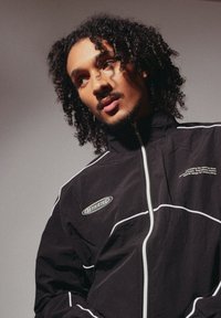 Μαύρο τζάκετ με φερμουάρ και λευκές λεπτομέρειες, κατασκευασμένο από ελαφρύ υλικό. Διαθέτει λογότυπο στο αριστερό στήθος και κείμενο κατά μήκος του φερμουάρ.