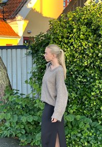 Béžový oversized svetr s žebrovanými manžetami, kombinovaný s černou sukní s bočním rozparkem, umístěný na pozadí bujné zeleně a dřevěného plotu.