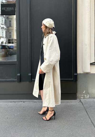 Abrigo ancho de color beige claro, vestido negro debajo, zapatos de tacón negro con tiras, y un pañuelo ligero con un patrón. Texturas suaves y tejido blando.