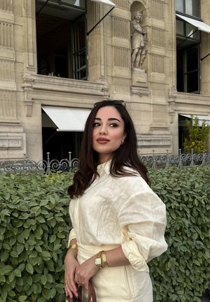 Camisa de lino crema con mangas remangadas, joyas doradas y un bolso marrón. El fondo presenta vegetación y detalles arquitectónicos ornamentados.