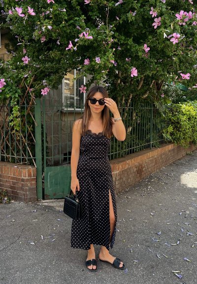 Vestido midi negro con lunares, detalles de encaje, abertura lateral, combinado con sandalias negras y un bolso pequeño. El fondo presenta vegetación y flores.