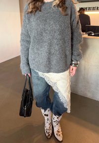 Personne portant un pull gris pelucheux, un tablier crème orné de dentelle par-dessus un jean, des bottes de cow-boy à pois, tenant un sac à main noir, à l'intérieur près d'un comptoir.