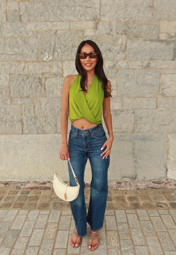 Femme portant un haut sans manches vert, un jean bleu, des sandales et des lunettes de soleil, tenant un sac à main blanc, debout contre un mur en pierre.