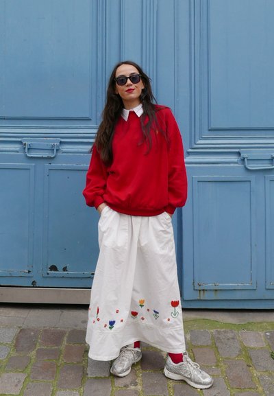 Sudadera roja con cuello blanco, combinada con una falda blanca que presenta bordados florales. Completada con zapatillas plateadas y gafas de sol oscuras.