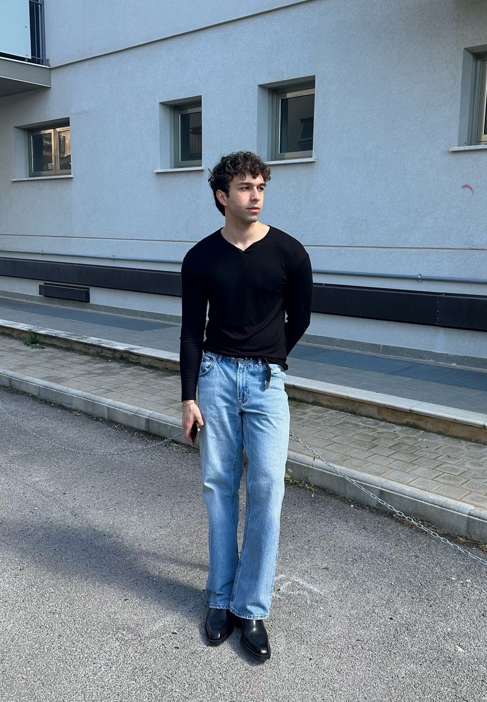 Black long-sleeve shirt and light blue jeans; wearing black ankle boots, standing on a street with a minimalistic backdrop.