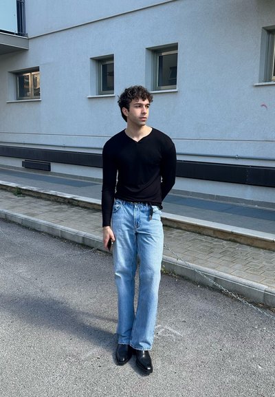 Camisa negra de manga larga y jeans azul claro; lleva botas negras hasta el tobillo, de pie en una calle con un fondo minimalista.