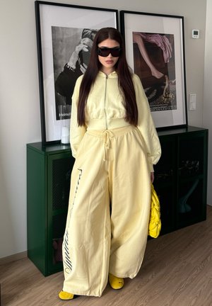 Mujer con cabello largo y oscuro, lleva pantalones amarillos anchos y sudadera con capucha y cremallera, gafas de sol negras, sostiene una bolsa amarilla, está de pie dentro de una habitación junto a un armario verde.