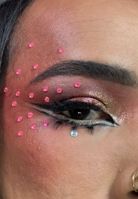 Primer plano de un ojo con sombra de ojos dorada y rosa brillante, delineado con lápiz negro, con pedrería rosa y un adorno de gema transparente.